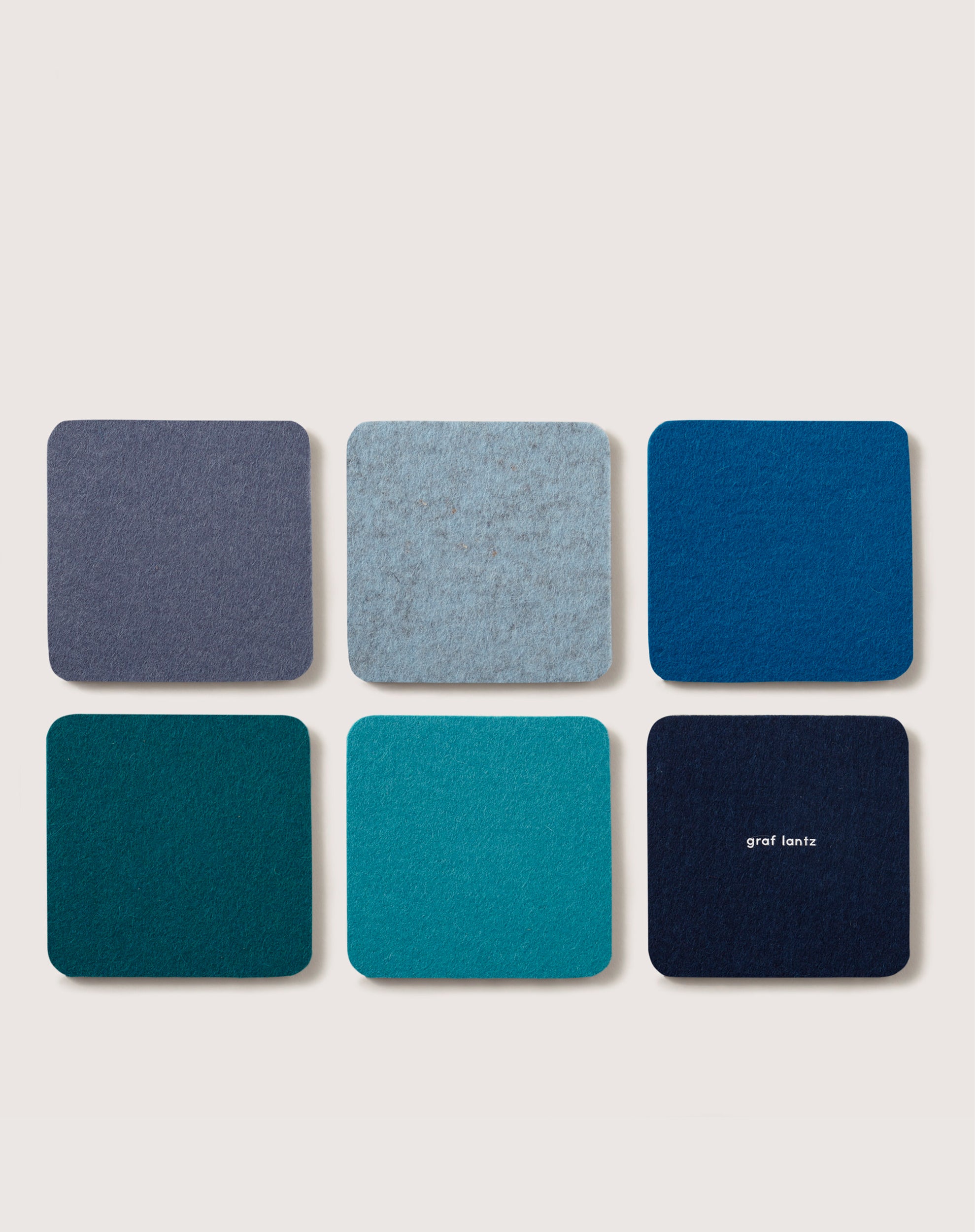 Six eco-friendly coasters made from German Merino wool are arranged in blue and teal hues. The bottom right coaster features the brand name graf lantz on a light, neutral background.