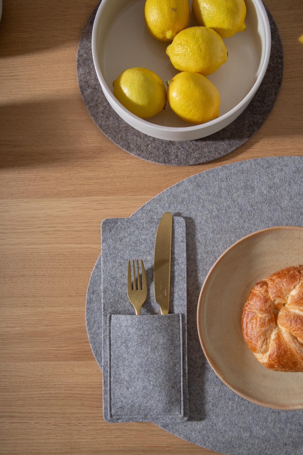 A bowl of lemons, a croissant on a plate, and cutlery are elegantly arranged on premium Merino wool placemats from graf lantz. The gray felt mats provide a designer-inspired look, enhancing the warmth of the wooden table setting.