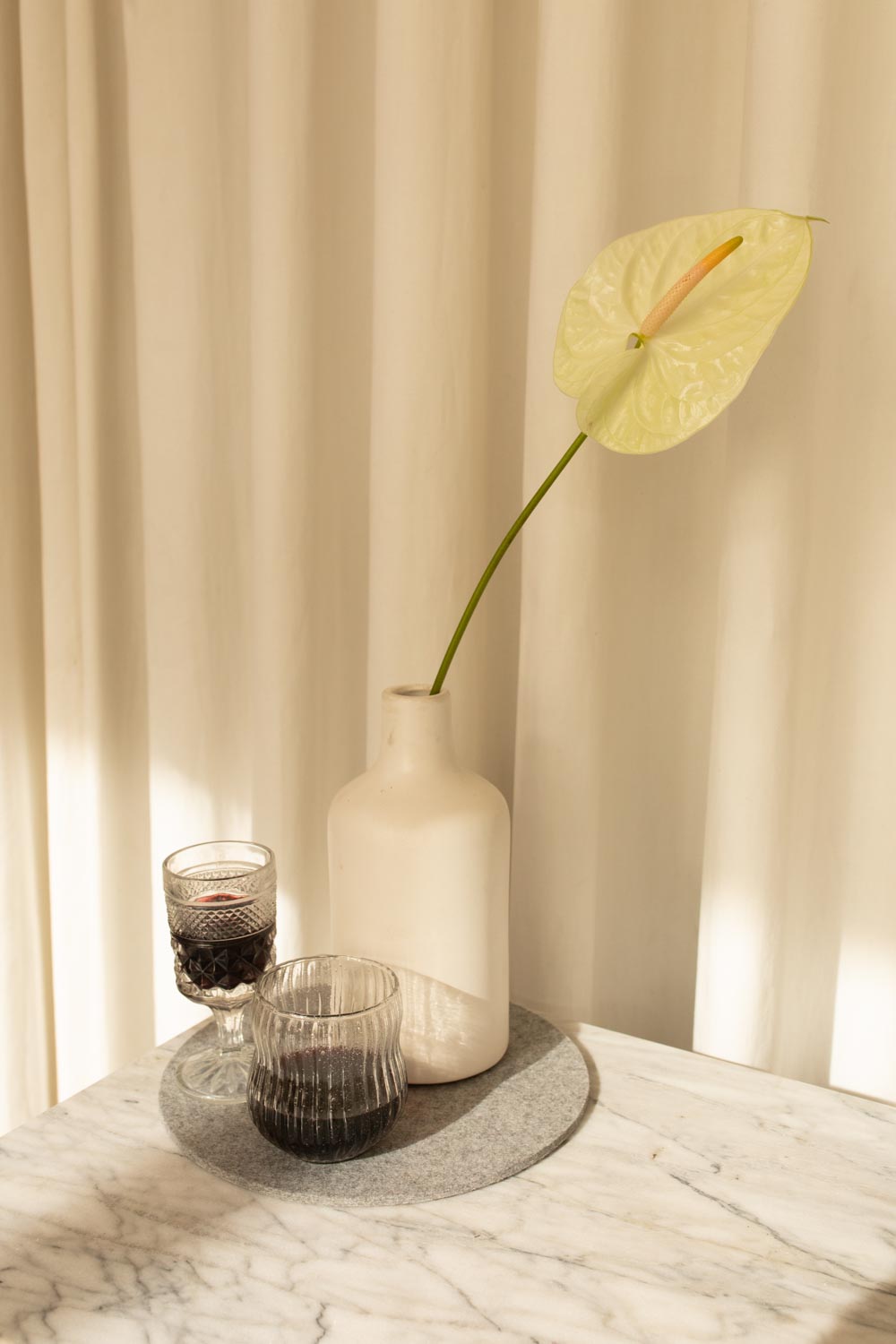A white vase with a pale green anthurium on marble is flanked by two glasses of dark red wine. Beige curtains in the background, softly lit by natural light, echo the gentle elegance of Merino wools drape, capturing a touch of graf lantz sophistication.