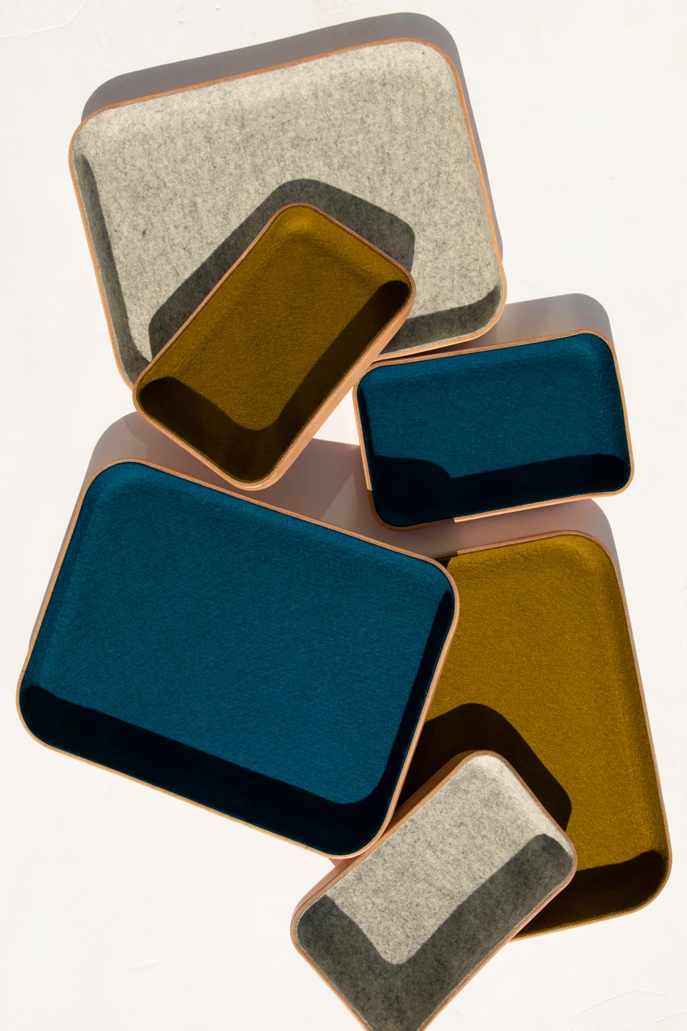 Five kawabon trays from graf lantz, in different sizes, rest on a light surface. They have Merino wool interiors in gray, dark blue, and mustard yellow hues with light brown borders. Sunlight dances over them, emphasizing their textures.