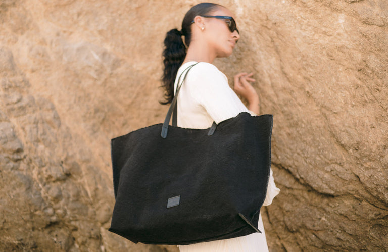 A person wearing sunglasses carries a large black tote bag in front of a rocky background. The person has long, dark hair pulled back and is wearing a light-colored outfit.