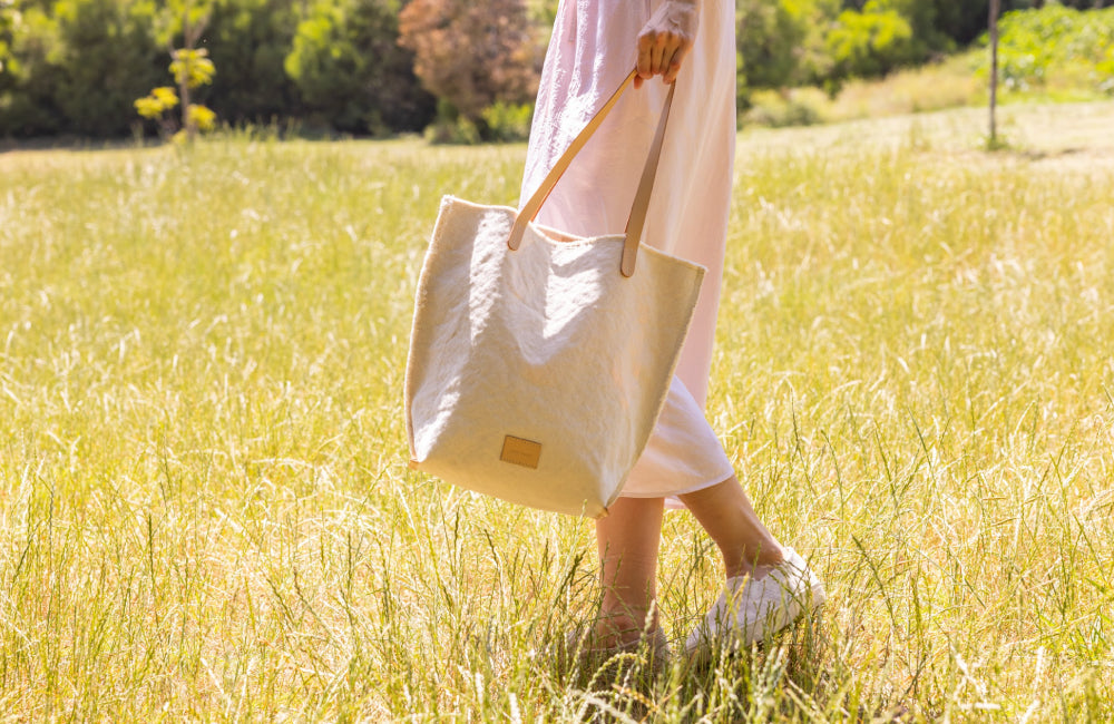 A person in a light pink dress stands in a sunny field, holding a large beige tote bag. Their face is not visible, and the grass is tall and green around them.