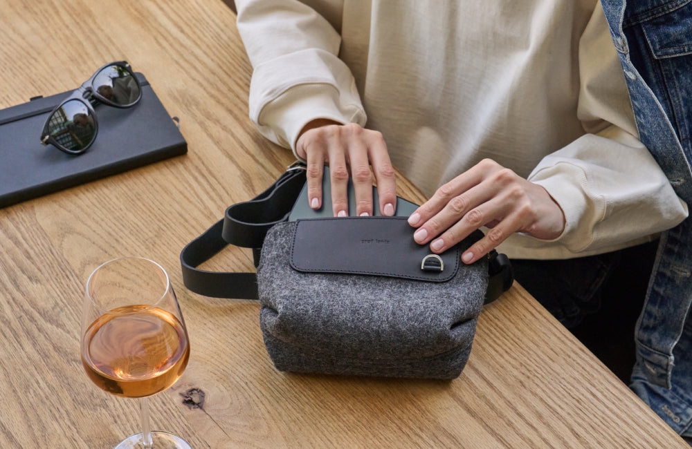 A person in a light sweatshirt opens a gray crossbody bag on a wooden table with a glass of rosé wine, sunglasses, and a closed laptop nearby.