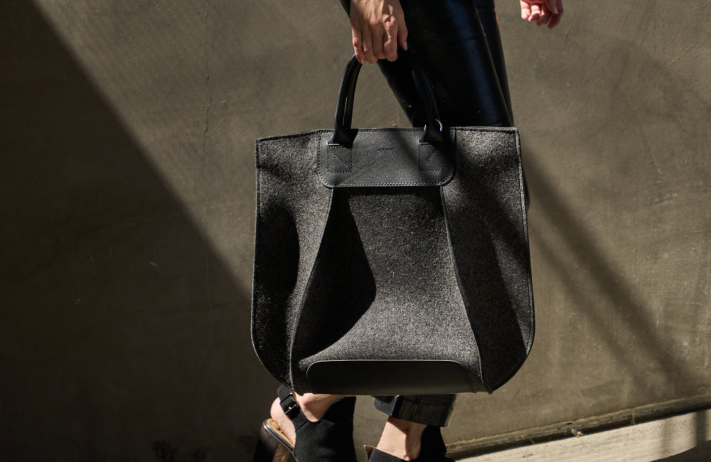 A person carries a large, structured black handbag with short handles. They are wearing black pants and open-toe heeled sandals, walking in sunlight against a textured wall.