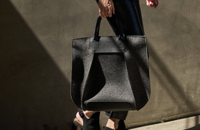 A person carries a large, structured black handbag with short handles. They are wearing black pants and open-toe heeled sandals, walking in sunlight against a textured wall.