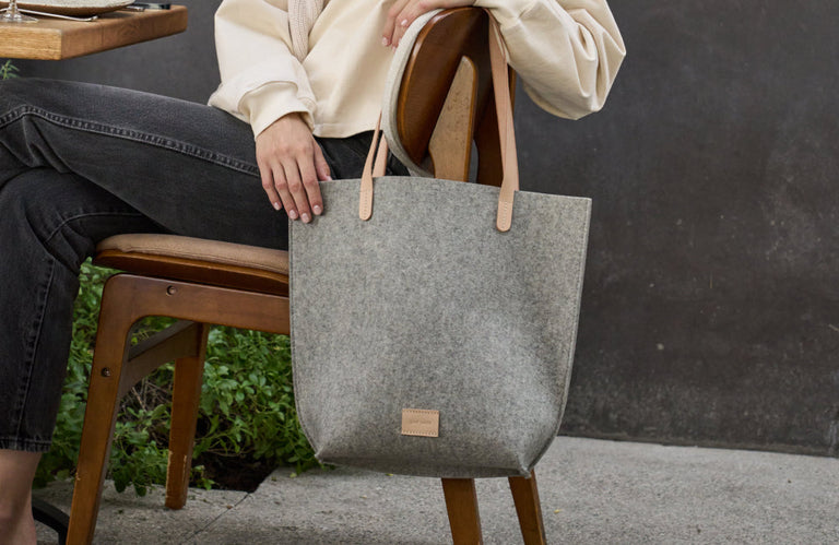A person in a cream sweater and dark jeans sits on a wooden chair, holding a large gray felt tote bag with tan handles. The background includes greenery and a dark wall.