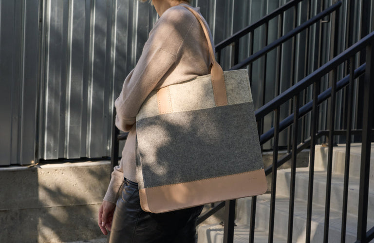 Person wearing a beige sweater and black pants stands outdoors holding a large, two-tone tote bag made of gray felt and tan leather, with stairs and a metal fence in the background.