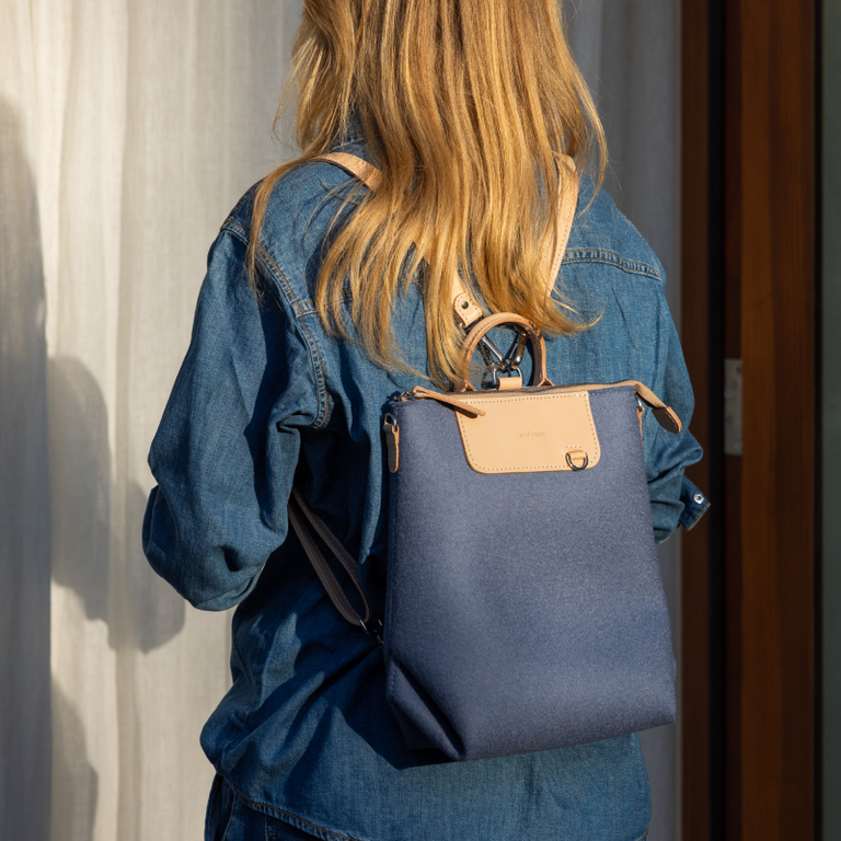 A person with long blonde hair wears a blue denim shirt and a blue backpack with a tan flap, standing in sunlight near a curtain and a wooden door.
