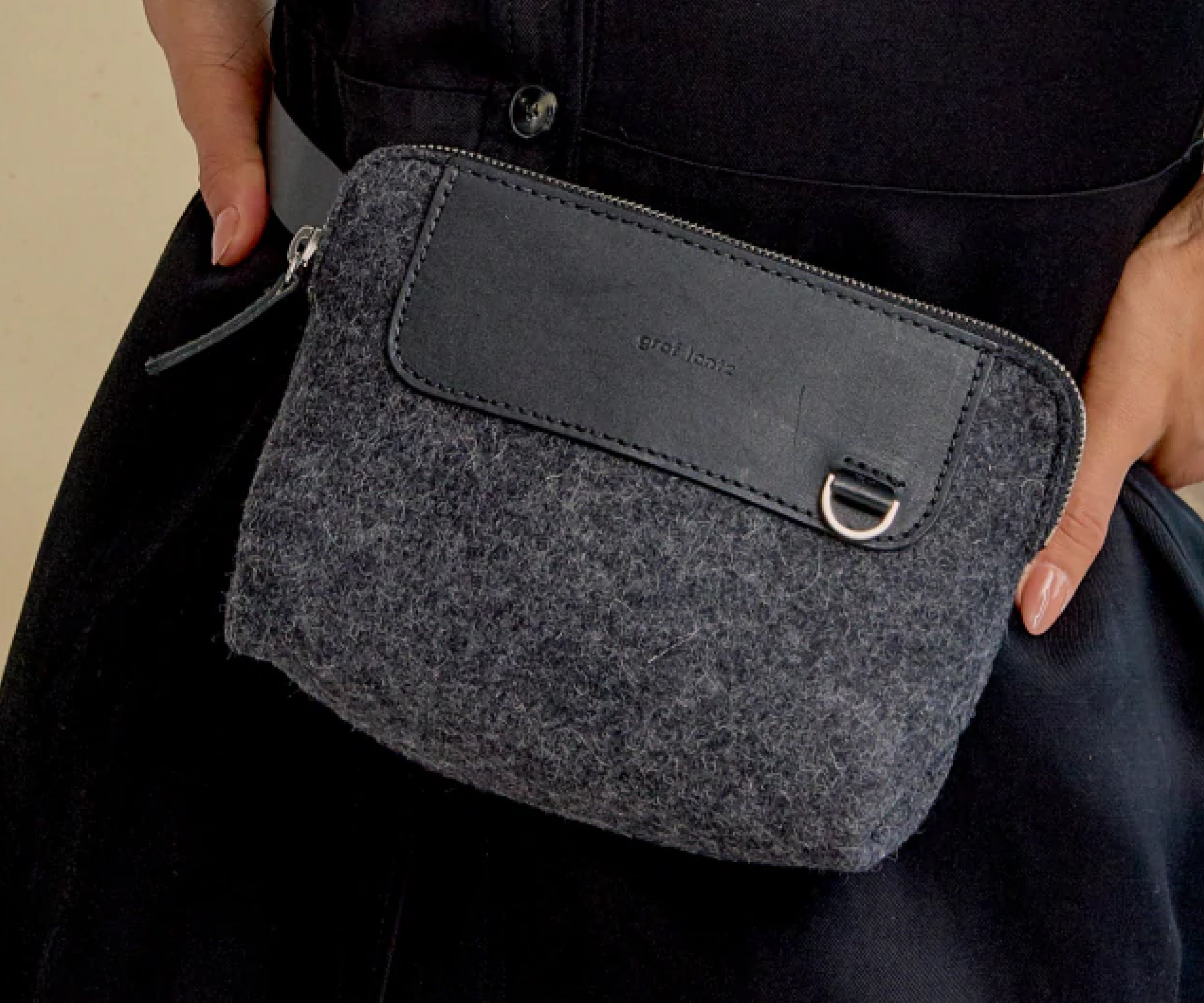 A person wearing dark clothing holds a small, gray felt pouch with a black leather accent, zipper closure, and a metal ring on the front.