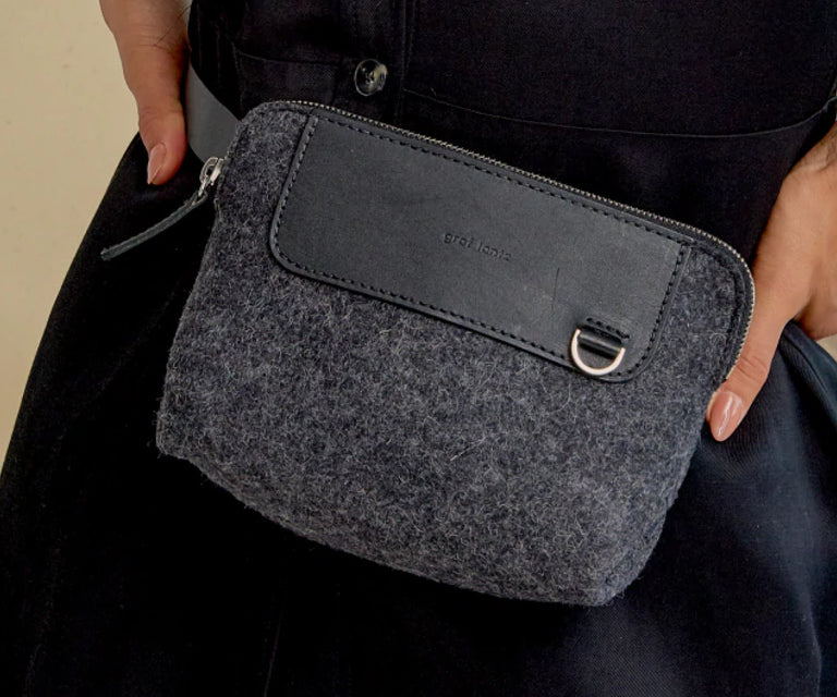 A person wearing dark clothing holds a small, gray felt pouch with a black leather accent, zipper closure, and a metal ring on the front.