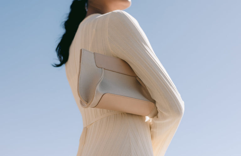 A woman in a light, long-sleeved dress holds a beige clutch purse against her side, standing against a clear blue sky. Her face is out of frame, and her dark hair is tied back.