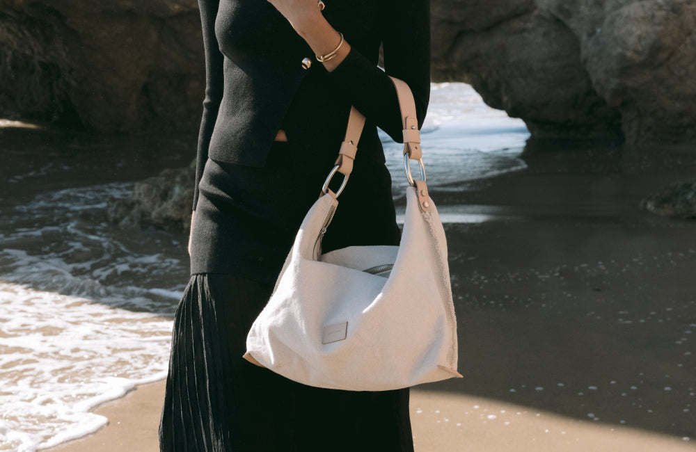 A person wearing a black outfit stands on a beach, holding a light-colored shoulder bag. Waves and rocks are visible in the background.
