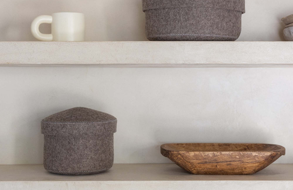 Two beige shelves display a white mug, two round gray felt containers with lids, and a shallow wooden bowl against a plain, light-colored wall.