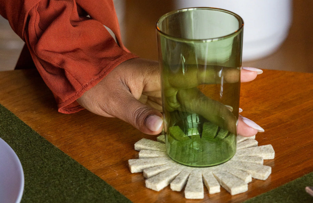 A person with a rust-colored sleeve places a green glass on a light-colored, sun-shaped coaster on a wooden table.