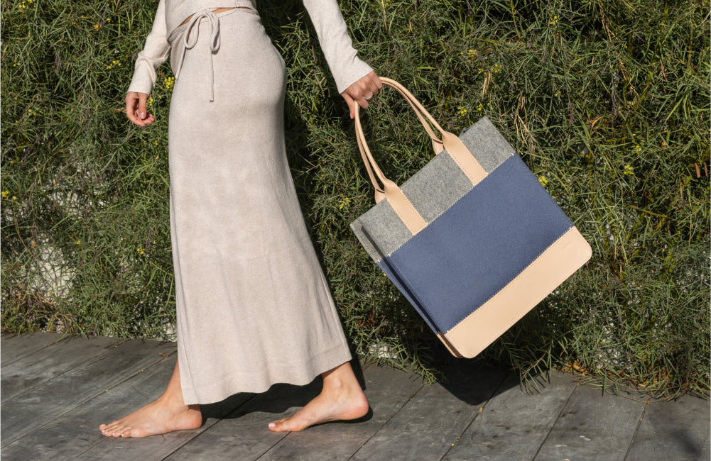 A barefoot person in a beige dress walks on a wooden path, holding a large tote bag with beige, gray, and navy blue color blocks, in front of green foliage.