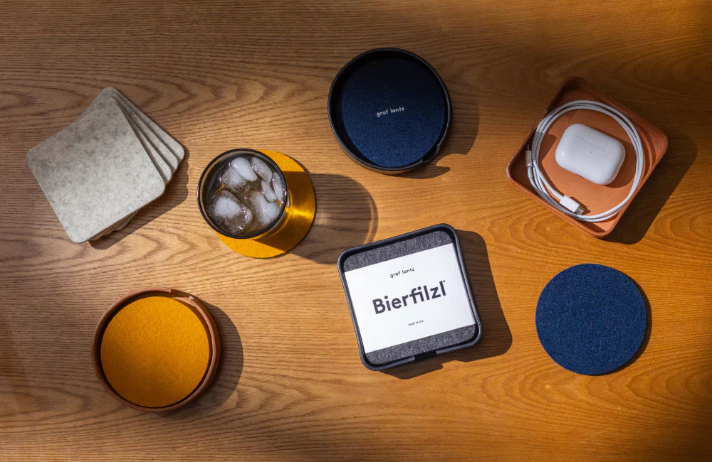 A wooden table with felt coasters, a glass of iced drink, a square container labeled Bierfilzl, a set of stacked coasters, a tray with a charging cable and AirPods, all in sunlight.