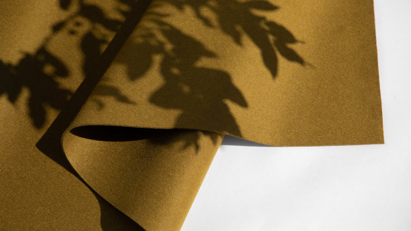 A piece of mustard yellow fabric lies on a white surface with the shadow of leafy branches cast across it.