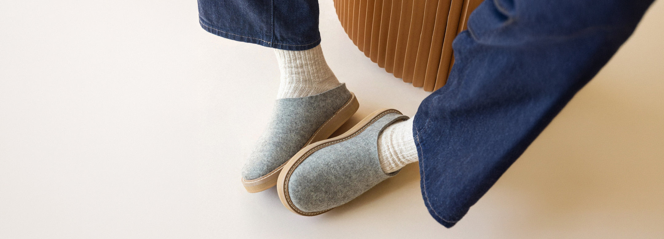 A person wearing cream socks and gray felt slip-on clogs with tan soles, paired with blue jeans, sitting on a light-colored surface.