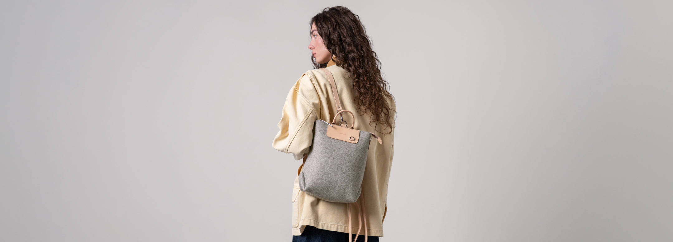 A person with long, light brown hair wearing a denim jacket is seen from behind, carrying a stylish blue and tan backpack outdoors in soft, natural sunlight.