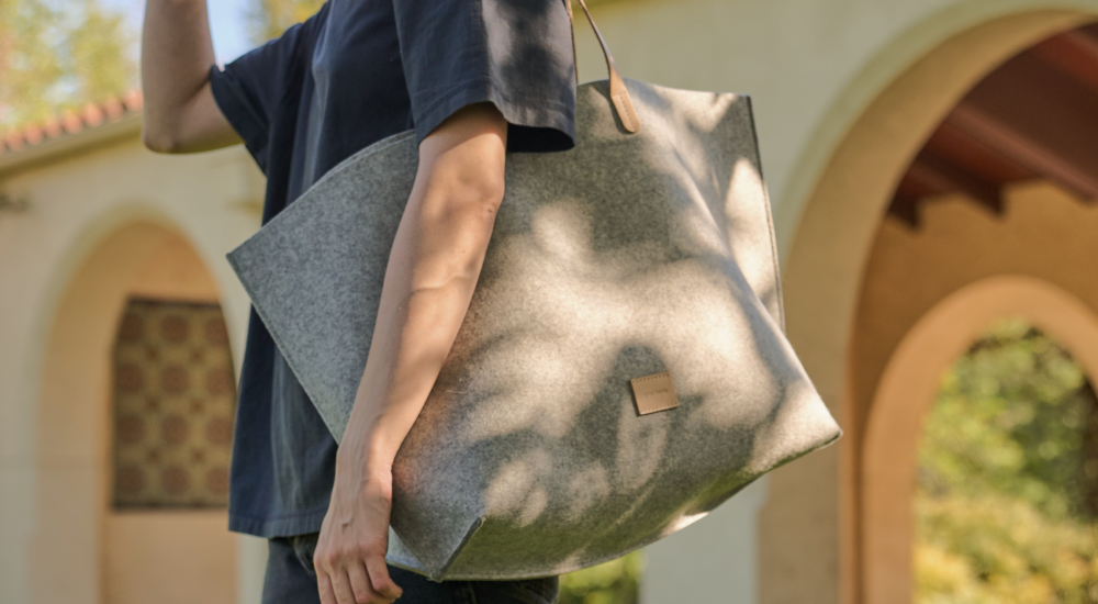 A person walking with a large, stylish black tote bag with geometric design, wearing black pants and wooden-heeled sandals. Sunlight casts shadows on a textured wall in the background.