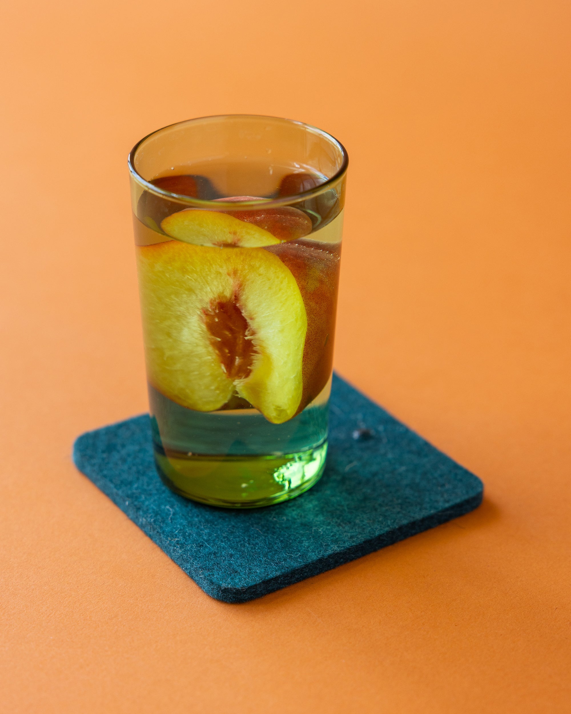 A glass of water with peach slices inside sits on a blue coaster against an orange background.
