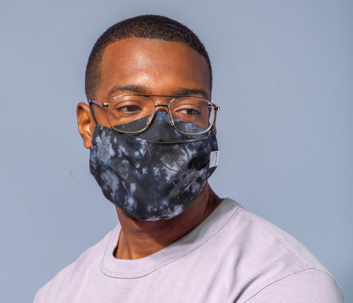 A man wearing glasses and a black-and-gray tie-dye face mask looks slightly to the side. He is dressed in a light gray shirt and poses against a plain light blue background.