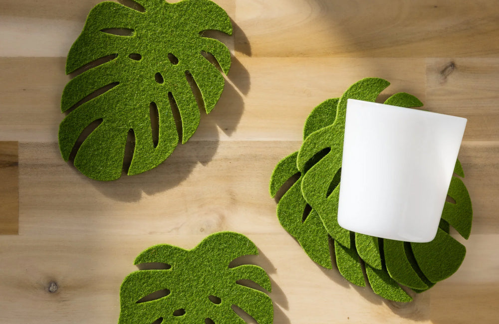 A white cup rests on green coasters shaped like monstera leaves, placed on a light wooden surface alongside more leaf-shaped coasters.