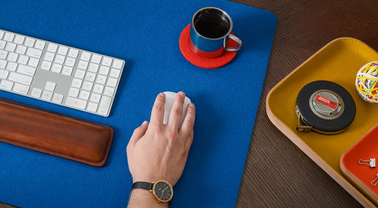 A laptop, mouse, notebook with glasses and coffee, and a tray with pens, clips, eraser, and earbuds are arranged neatly on a wooden desk.