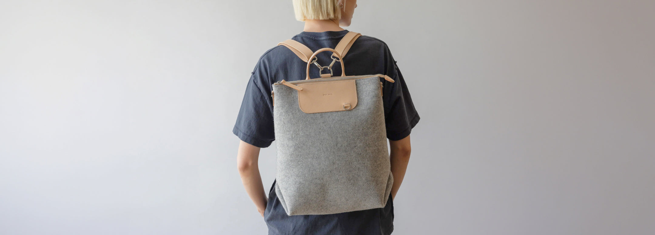 A person with short blonde hair, wearing a black shirt and dark pants, stands facing away, carrying a grey backpack with tan leather straps and a front pocket, against a plain light background.