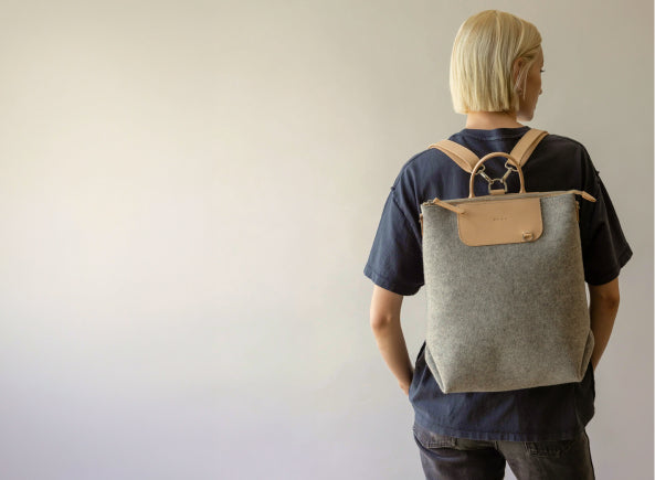 A person with short blonde hair, wearing a black shirt and dark pants, stands facing away, carrying a grey backpack with tan leather straps and a front pocket, against a plain light background.