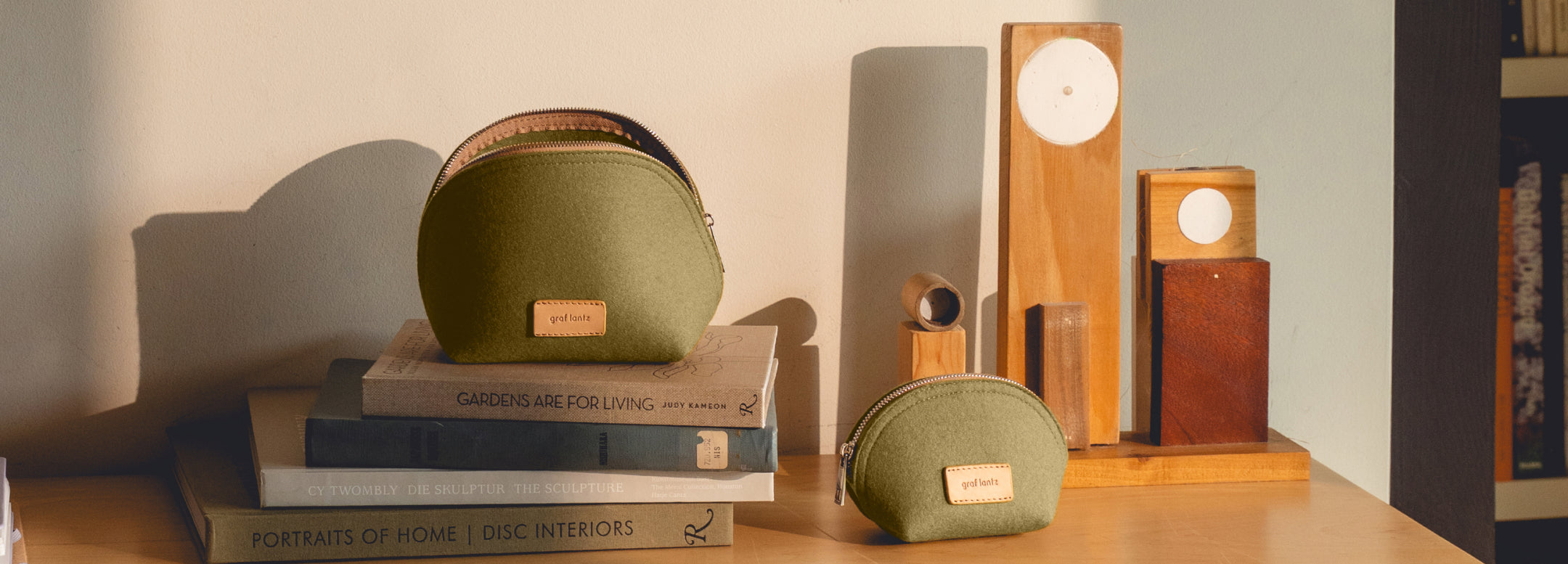 Two green zippered pouches sit on stacked books next to geometric wooden decor pieces on a sunlit wooden surface, with a bookshelf partially visible in the background.