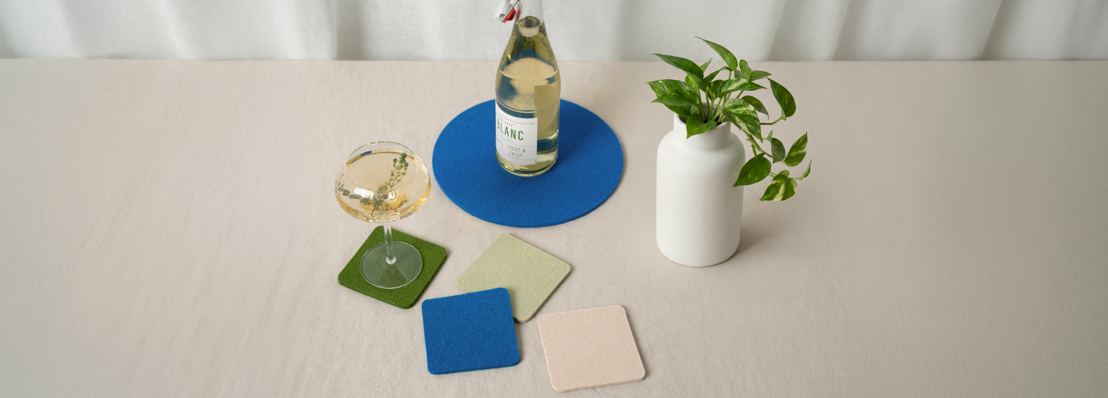A table with a wine glass, a bottle of white wine, colorful square coasters, and a small green plant in a white vase on a light tablecloth.