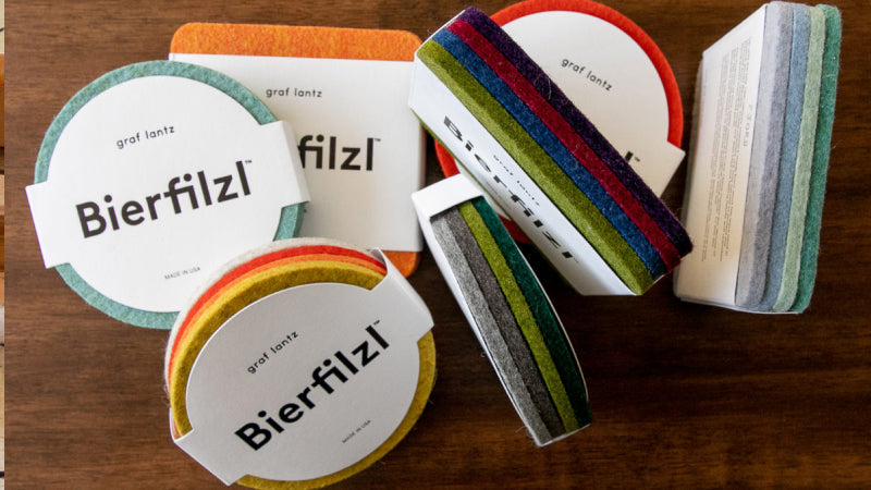 Felt coasters displayed on a wooden surface with a drink glass nearby. The coasters are in various colors, including blue, yellow, and gray, with a container labeled Bierfilzl at the center.