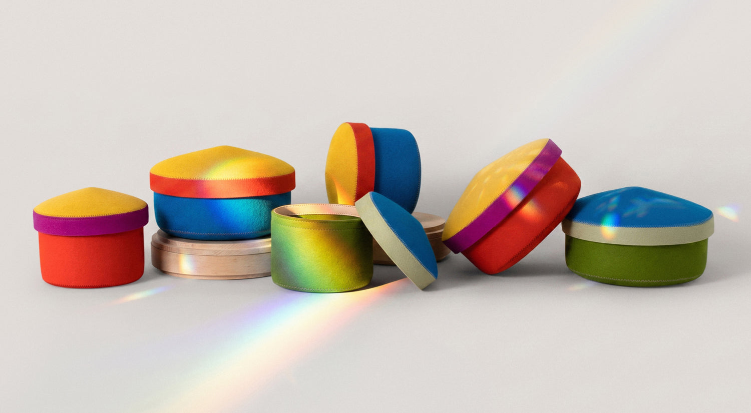 Colorful, round and heart-shaped containers with soft, fabric-like lids are arranged on a light surface. Rainbows from refracted light cross over the containers.