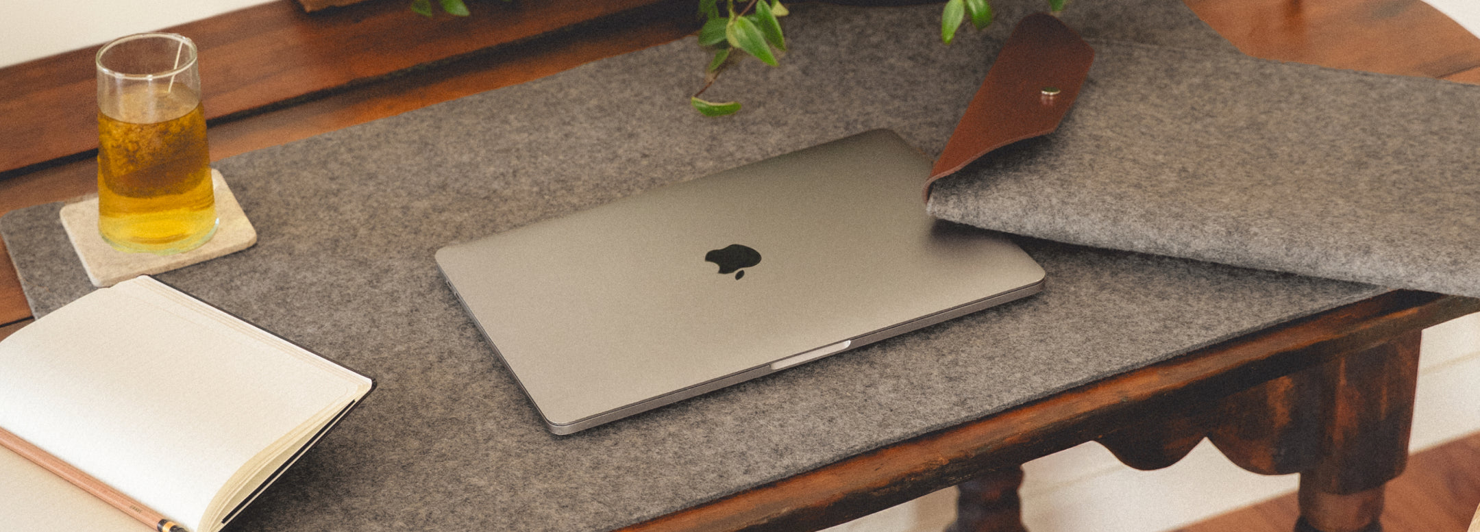 A laptop with a partially visible Apple logo sits on a scalloped mat on a wooden table. Nearby are a closed laptop sleeve and two pouches stacked on books. Soft shadows from tree branches dapple the scene.