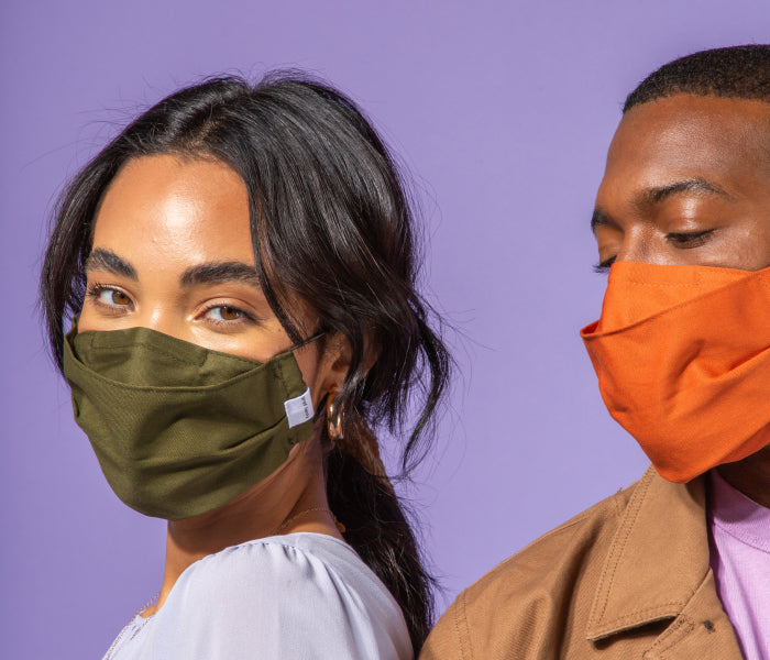 Two people wearing cloth face masks: the woman on the left wears an olive green mask and looks at the camera, while the man on the right wears an orange mask and looks downward. Purple background.