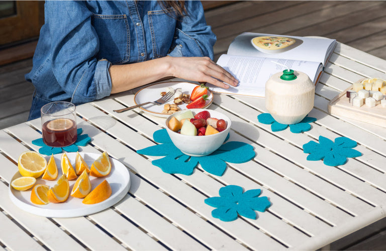 A person in a denim shirt sits at a table with fruit, a drink, a coconut, snacks, and an open cookbook. Blue flower-shaped mats decorate the white table.