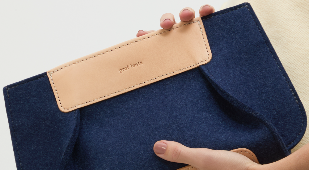 A hand holding a navy blue felt and beige leather clutch with the brand name graf lantz embossed on the leather flap.
