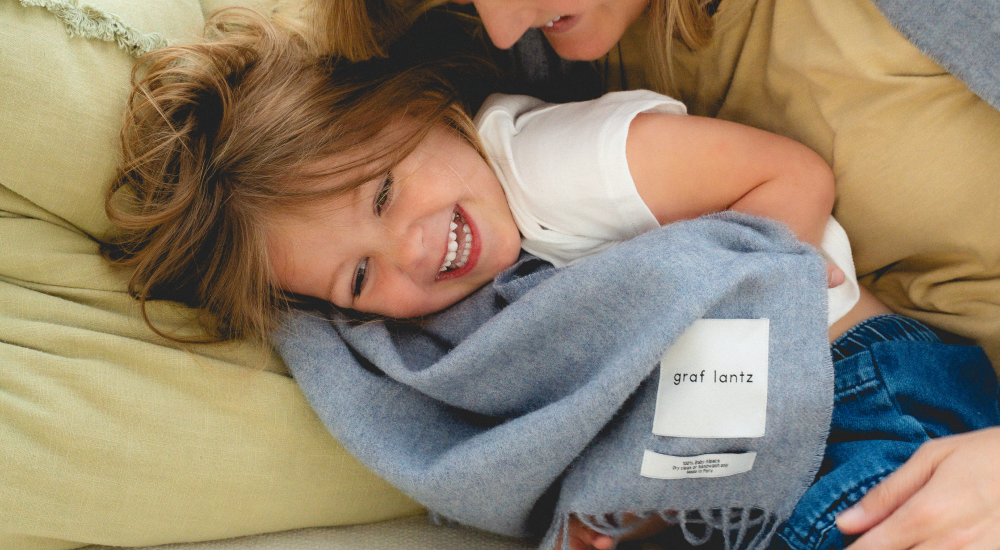 A smiling child lies on a couch, wrapped in a gray blanket labeled graf lantz, while being cuddled by an adult. The atmosphere is warm and cozy.