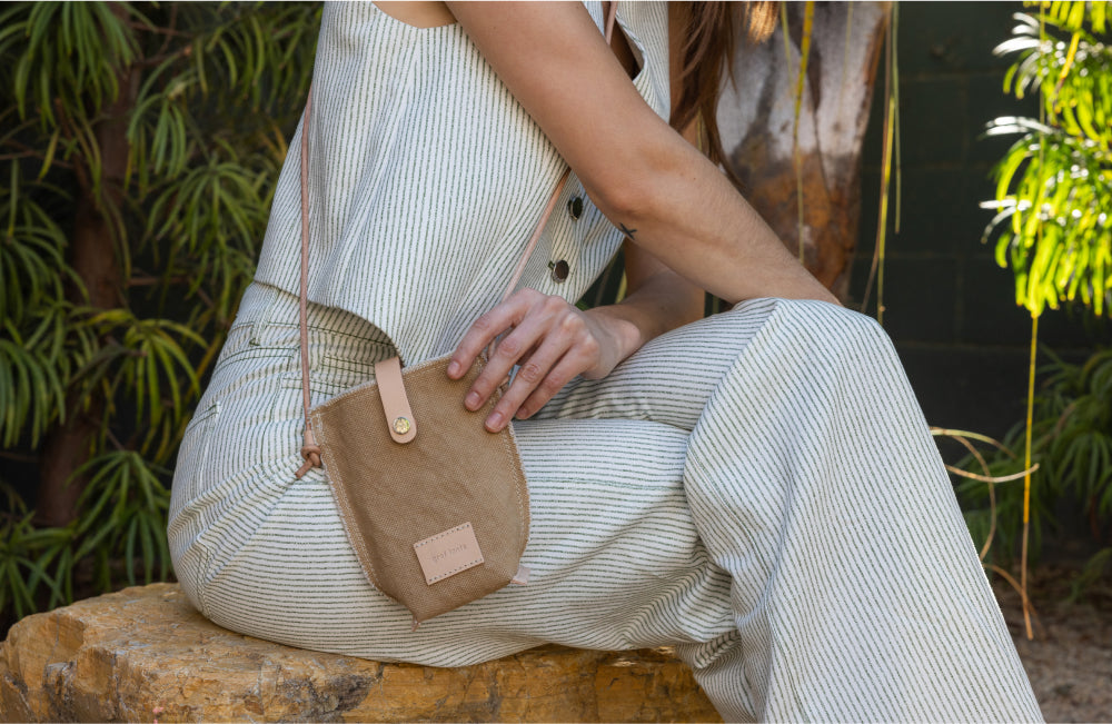 A person in a light-striped sleeveless jumpsuit sits on a rock outdoors, holding a small tan crossbody bag with a button clasp. Green plants fill the background.