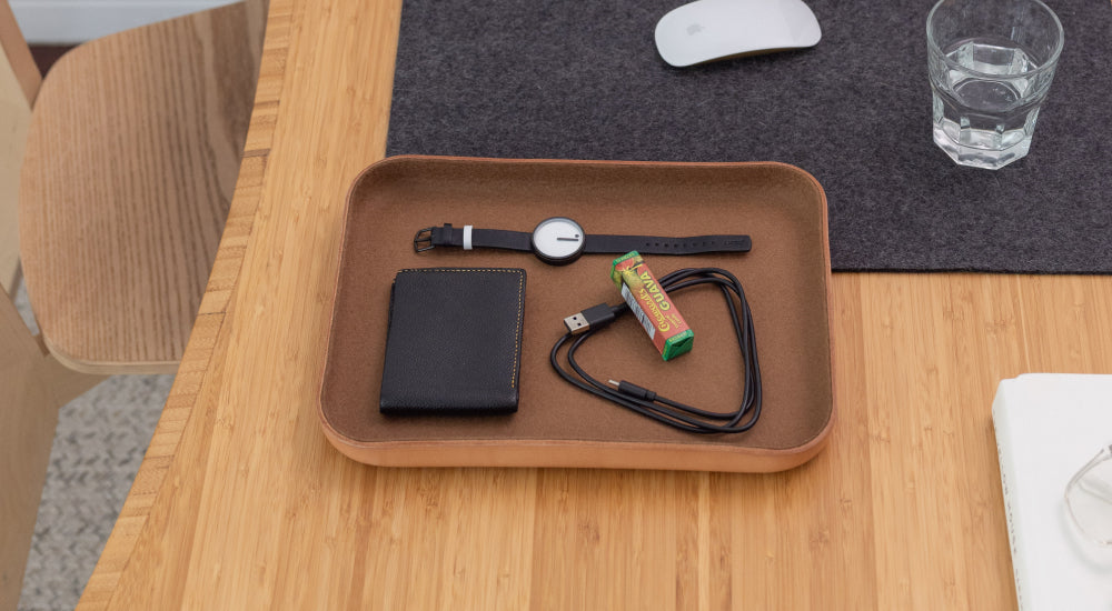 A leather tray on a wooden table holds a black wallet, a wristwatch, gum, and a charging cable. Nearby are a computer mouse, a glass of water, and a gray felt mat.