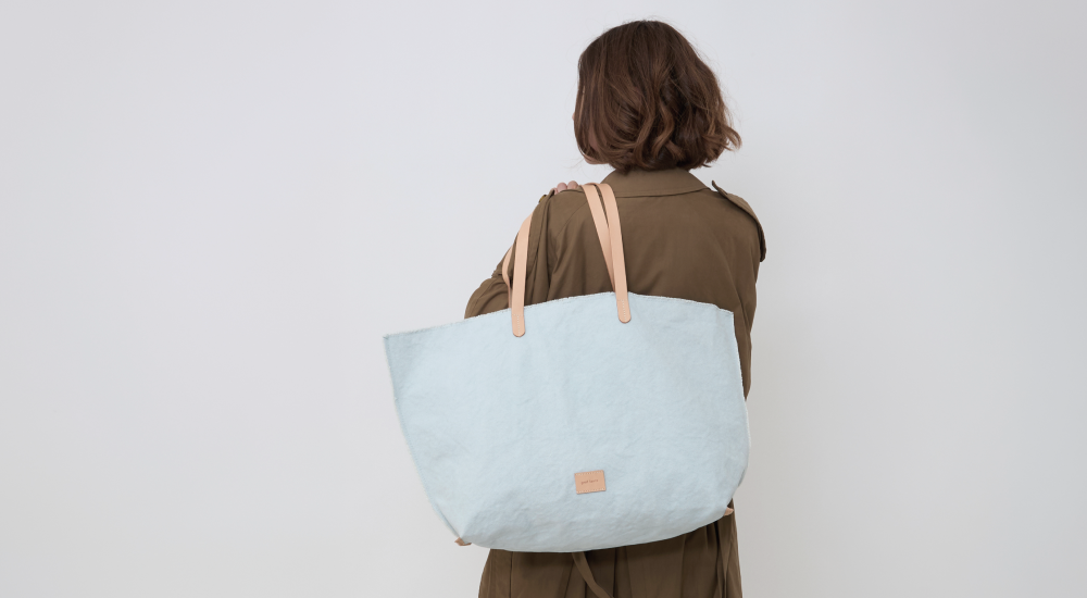 A person with shoulder-length brown hair, wearing a brown coat, faces away while carrying a large light blue tote bag with tan handles over their shoulder against a plain white background.