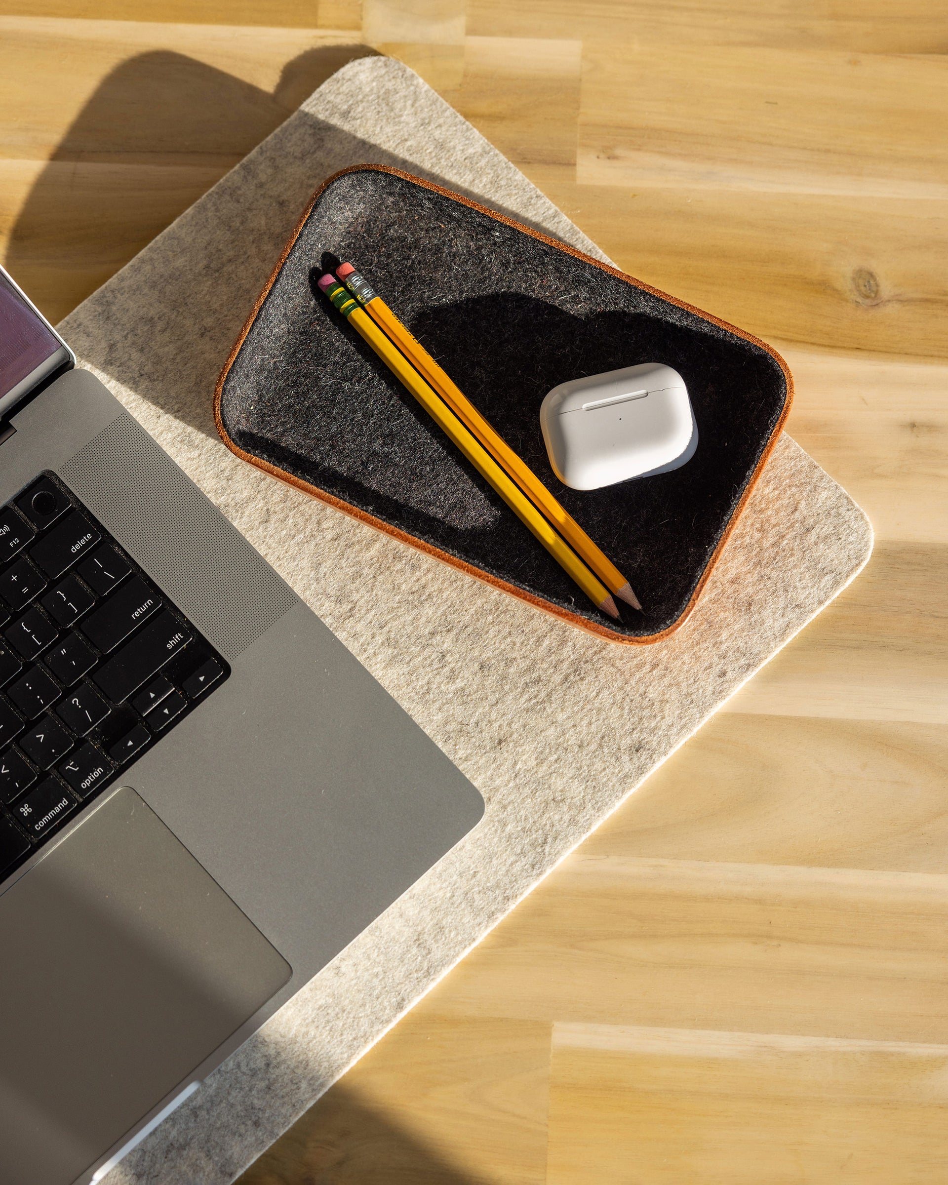 A laptop rests on a light wooden surface beside a graf lantz Merino wool catchall tray holding two yellow pencils and wireless earbuds in a white case, illuminated by natural sunlight.