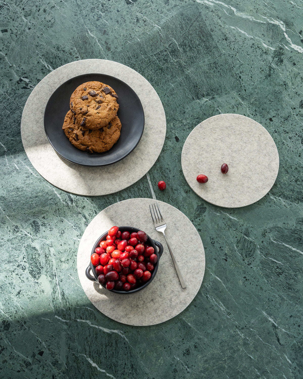 From above, a green marble table displays two round, beige placemats. One placemat holds a bowl of cranberries and a fork; the other features a black plate with chocolate chip cookies. Scattered cranberries add charm beside an Oeko-Tex® Certified trivet from graf lantz.