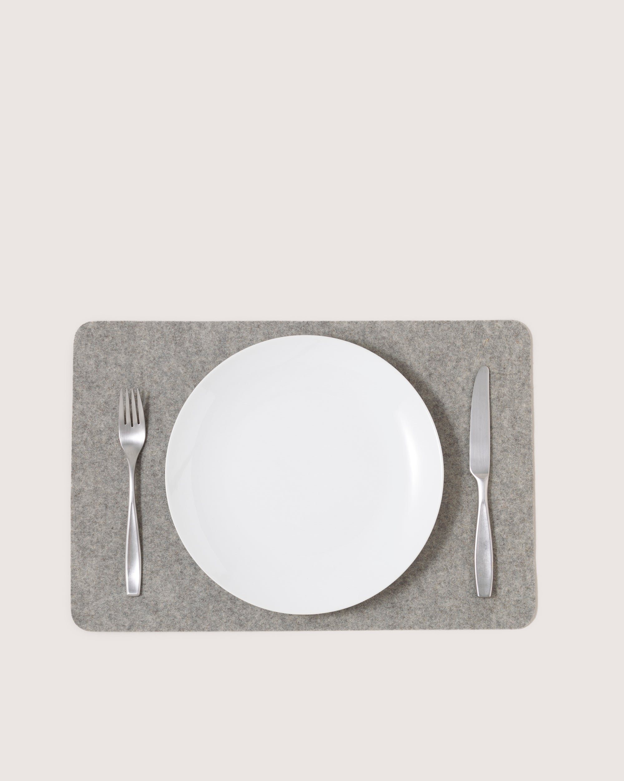 A white plate is centered on a heat-resistant merino wool placemat by graf lantz. A stainless steel fork rests to the left and a knife to the right, all elegantly set against a light gray background.