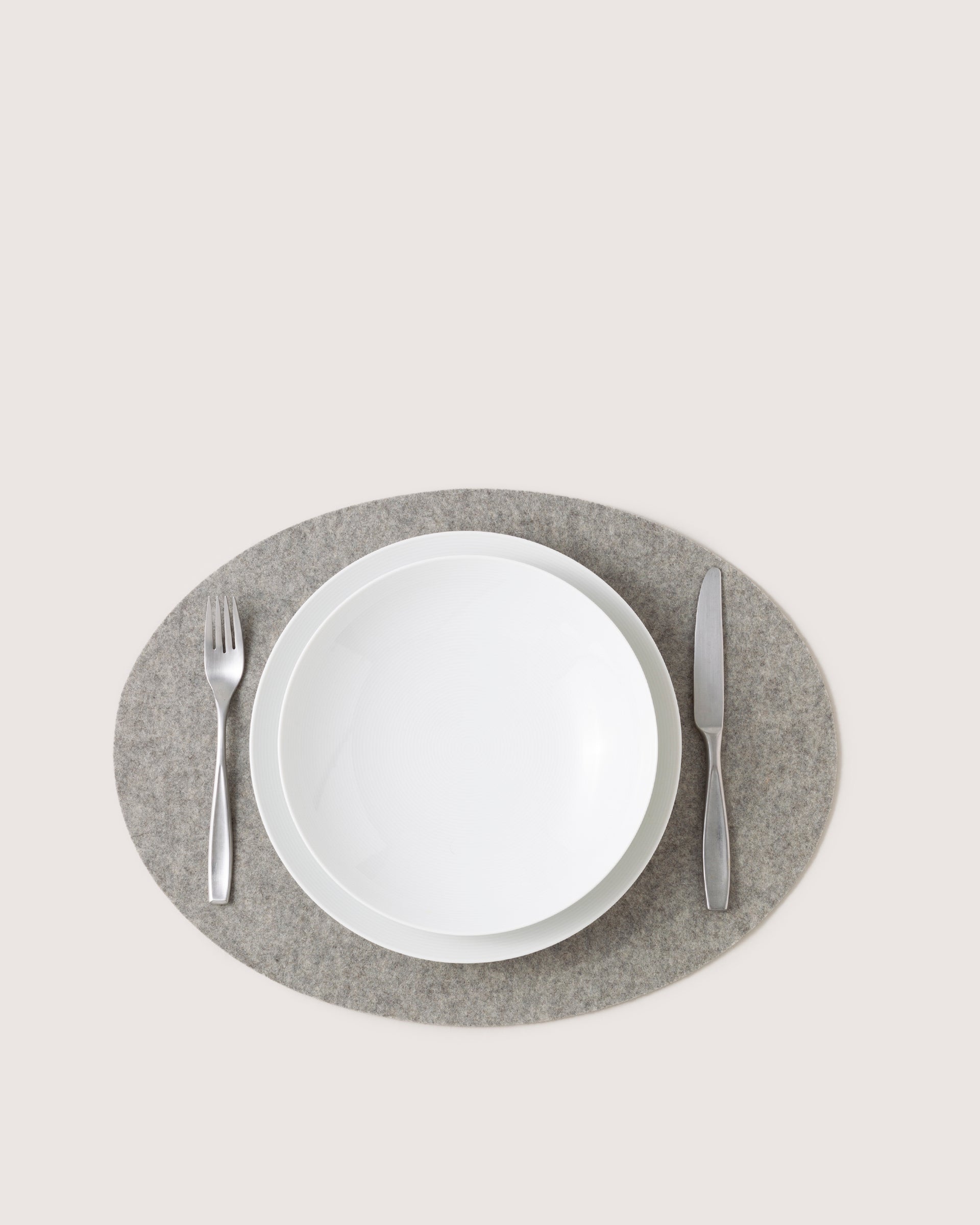 A white plate is centered on a premium graf lantz Merino wool placemat. A fork rests to the left and a knife to the right, both gleaming with metallic finishes. The heat-resistant backdrop remains light and plain, highlighting the refined setting.