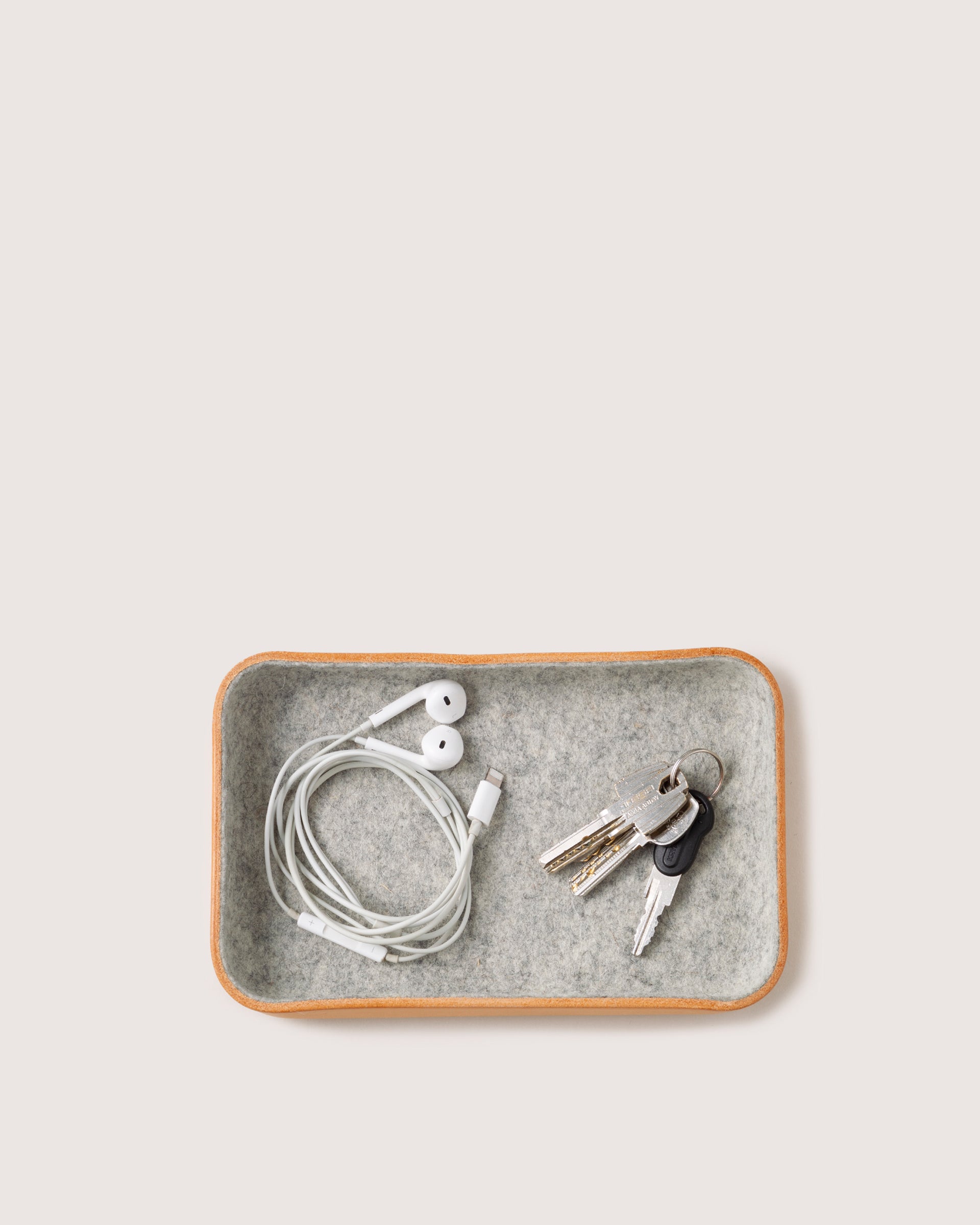 A graf lantz rectangular Kawabon Tray, crafted with impeccable design, neatly cradles coiled white earphones and keys adorned with a black keychain, against its soft, neutral backdrop that accentuates its elegance.