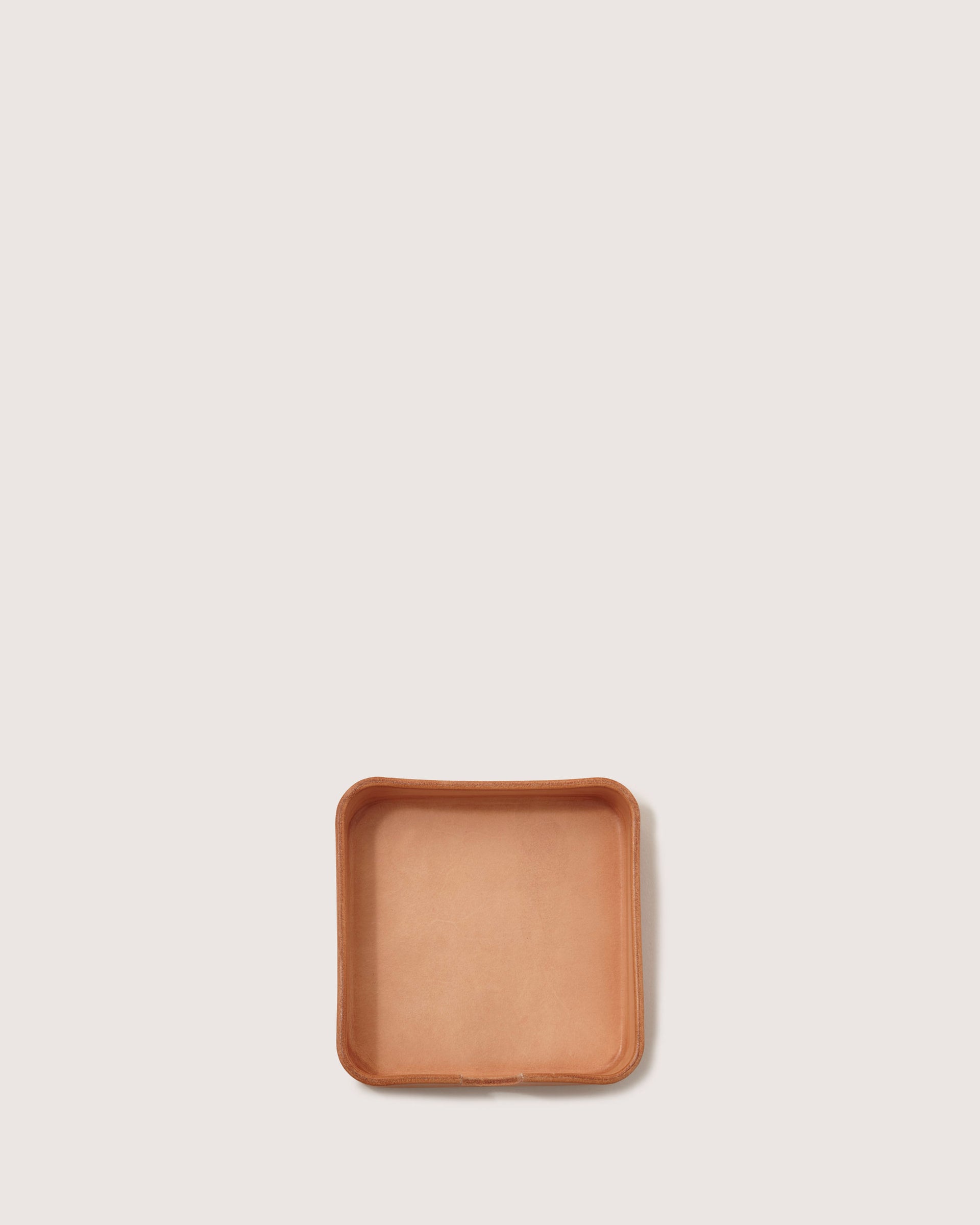 A small, square Vachetta leather tray from graf lantz with raised edges sits on a plain white backdrop. Its light tan color and smooth, slightly rounded corners exude elegance, making it a versatile organizer for keeping essentials neatly in place.