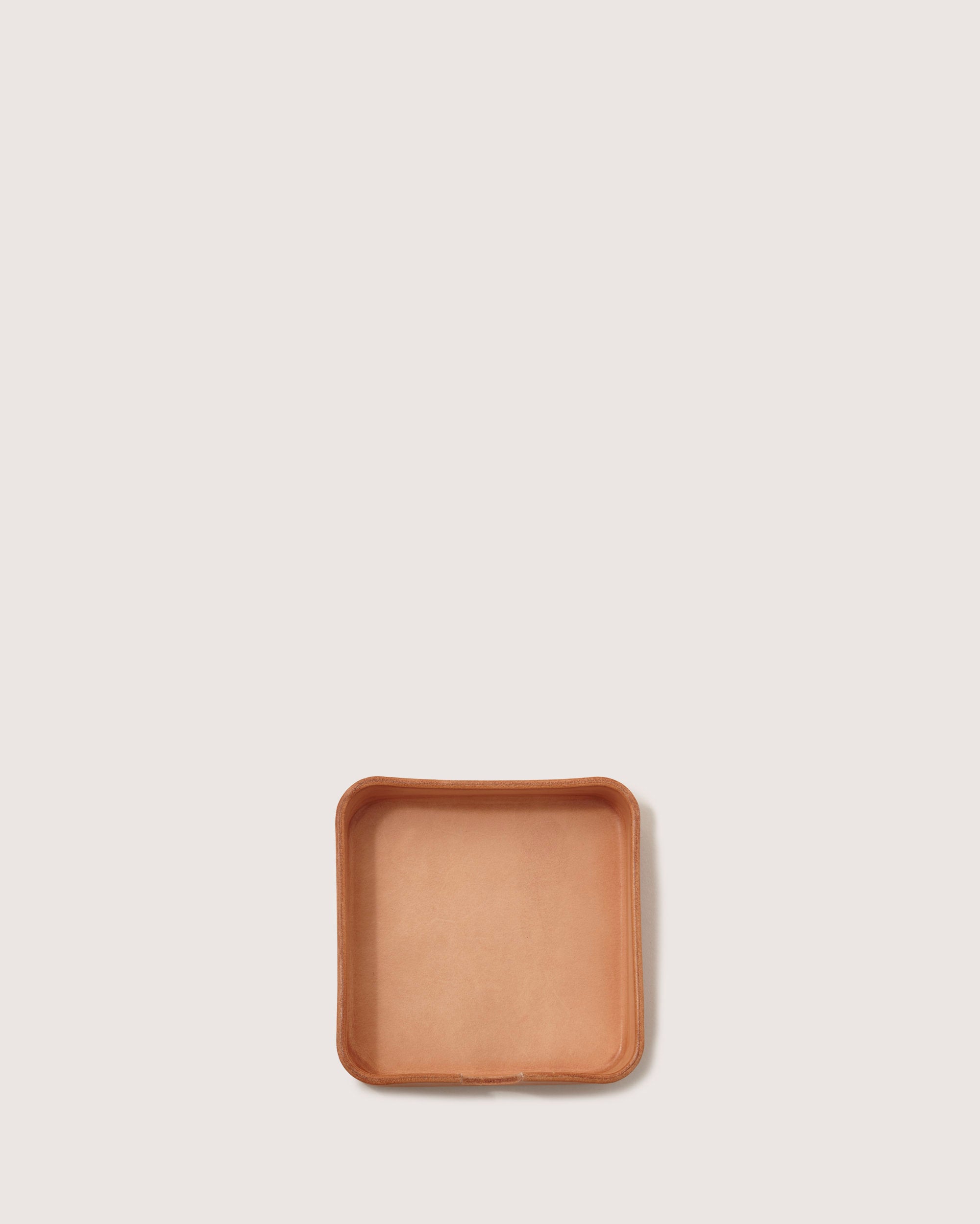 A small, square Vachetta leather tray from graf lantz with raised edges sits on a plain white backdrop. Its light tan color and smooth, slightly rounded corners exude elegance, making it a versatile organizer for keeping essentials neatly in place.
