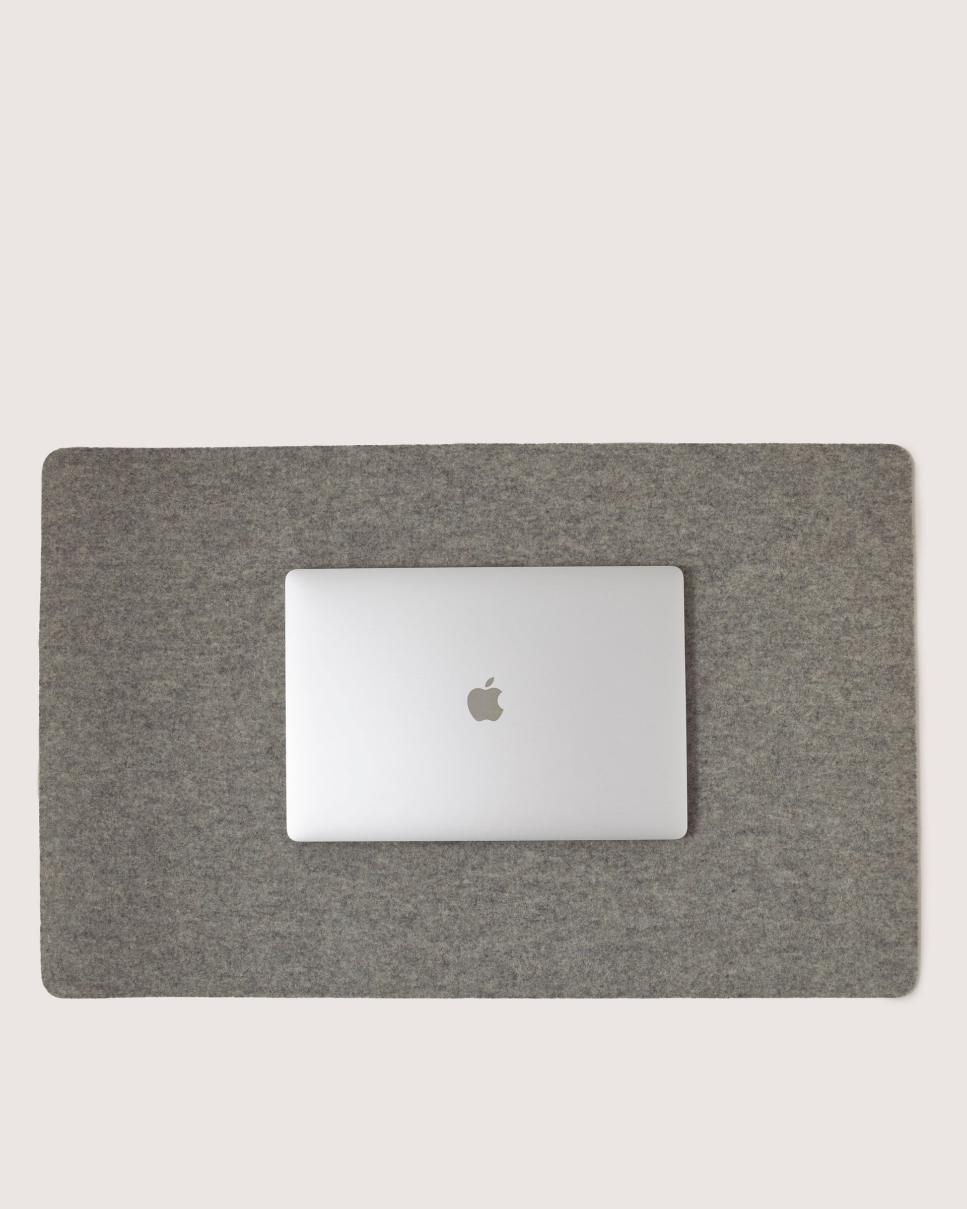 A closed silver laptop with an apple logo rests on a graf lantz Desk Pad crafted from luxurious Merino Wool. The plain background highlights this sleek workspace, emphasizing the elegant contrast between the laptop and the refined gray felt mat.